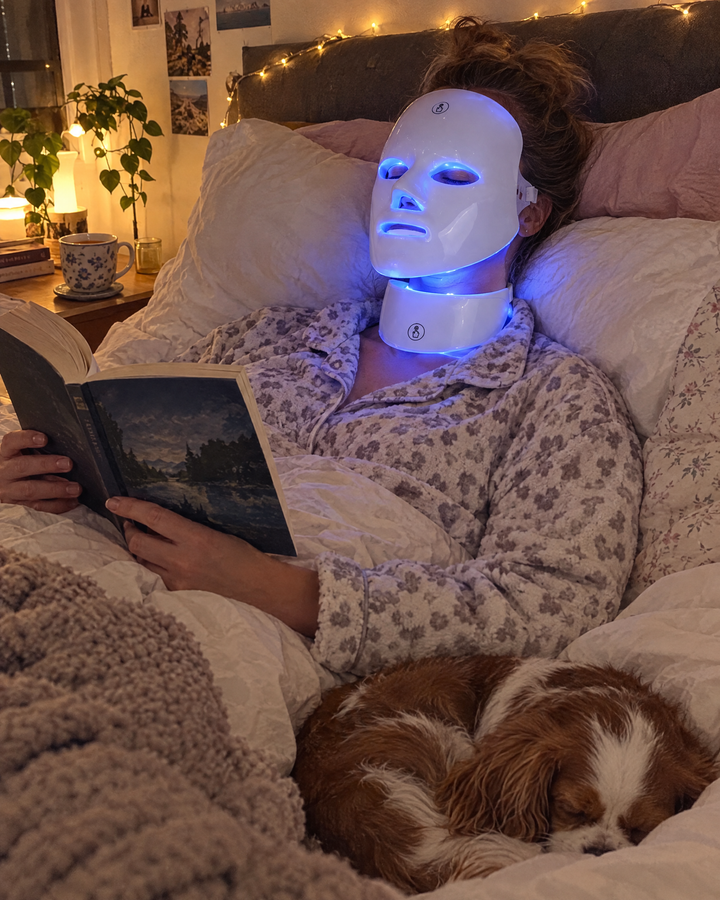 Woman reading in bed wearing the LED face and neck mask in blue light