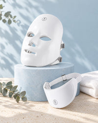 Himra LED face mask on a pale blue stone plinth with the neck piece, eucalyptus and a towel